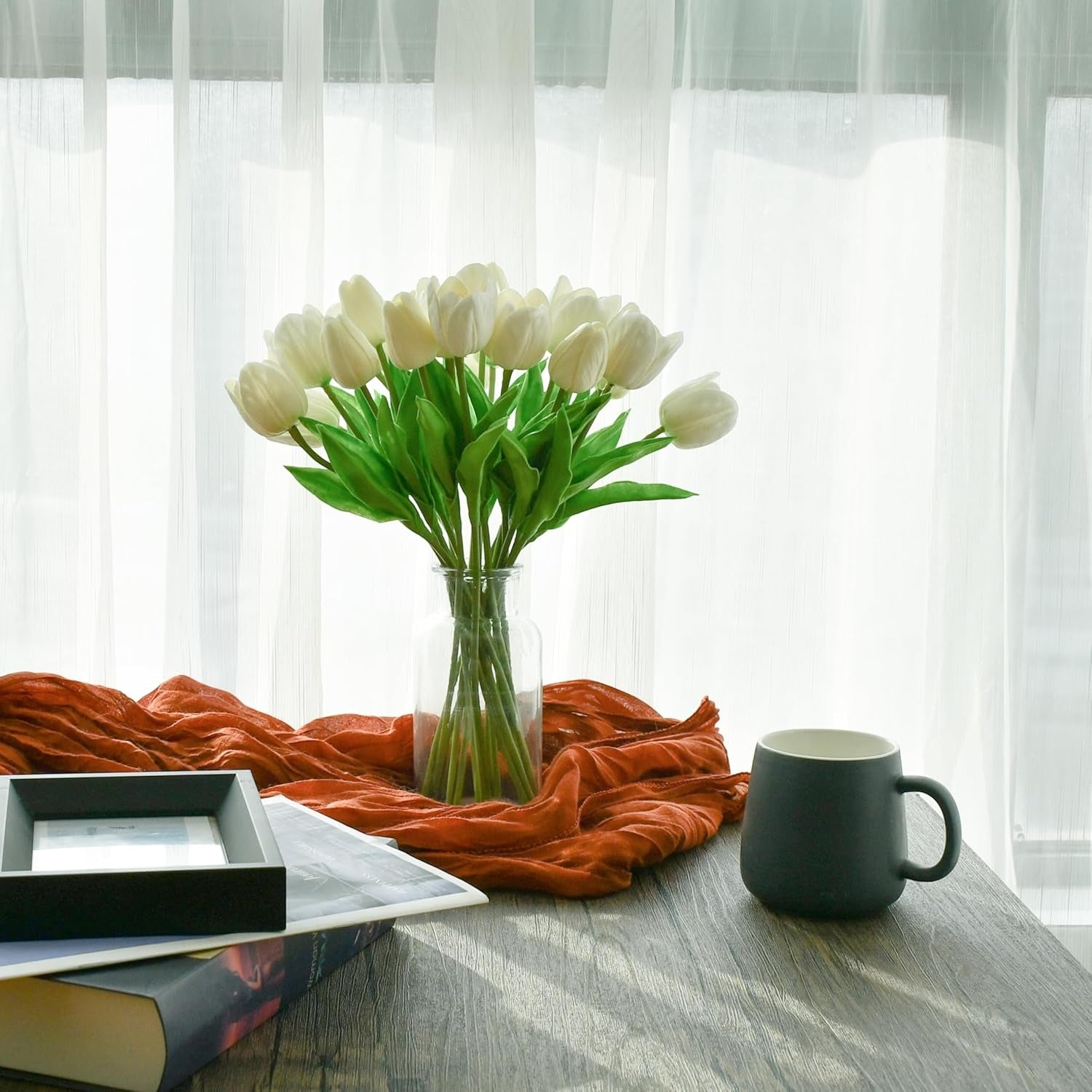 Bouquet of white tulips in a vase on a table with a mug and books, against a white curtain background.