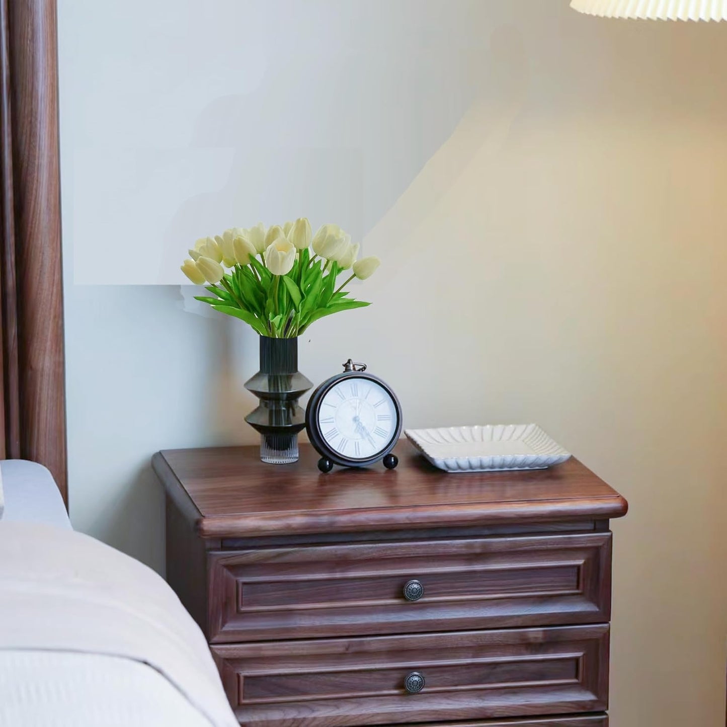 Wooden nightstand with a clock, vase of flowers, and plate on a blurred bedroom background