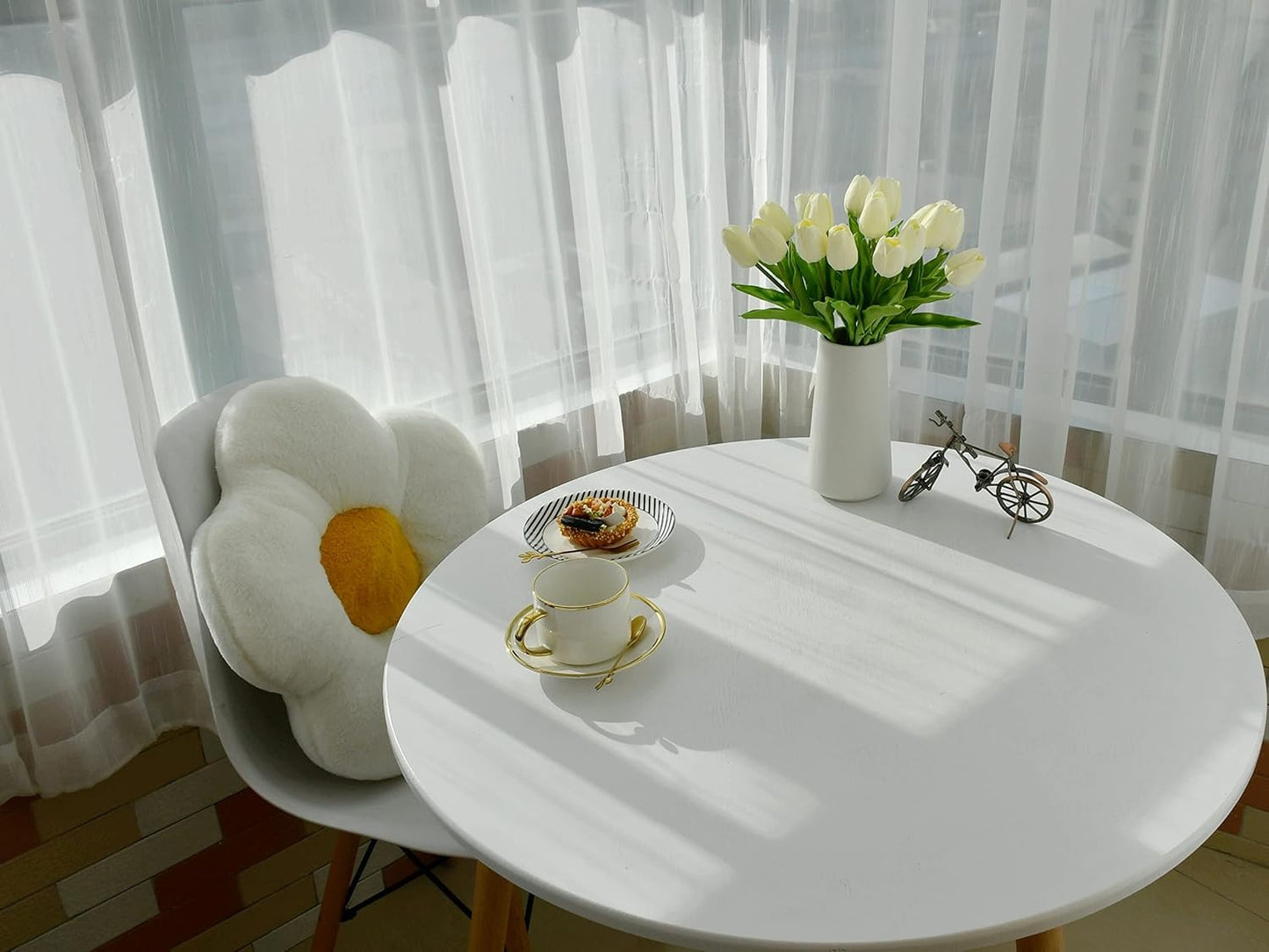 Round white table with a vase of white tulips, a teacup, and a small bicycle model in a bright room.