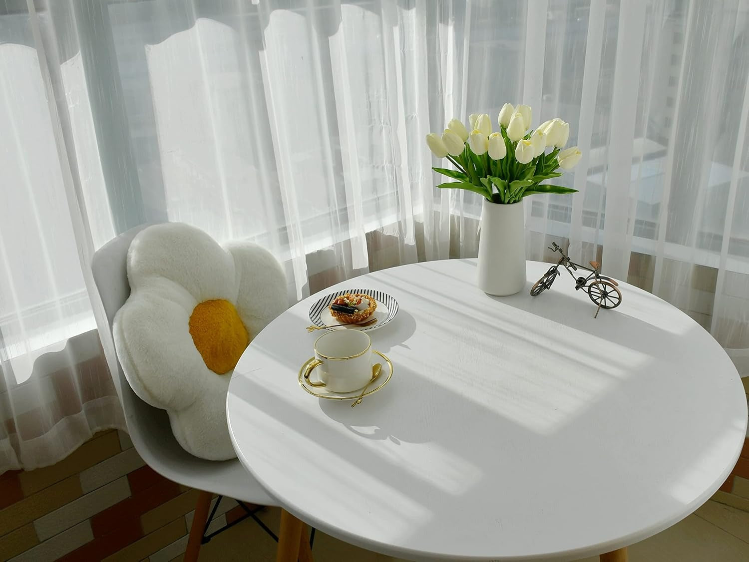Round white table with a vase of white tulips, a teacup, and a small bicycle model in a bright room.