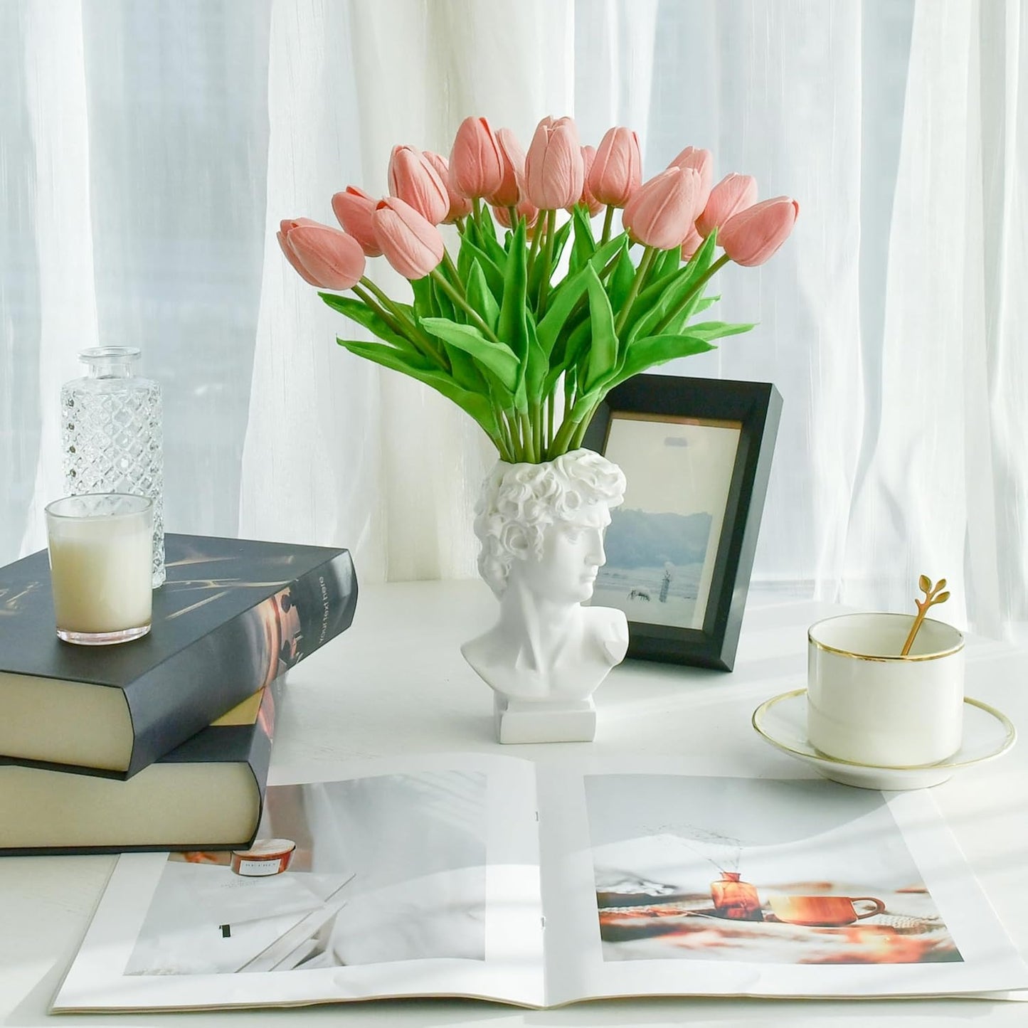 Pink tulips in a white vase on a table with books, a cup, and a candle.