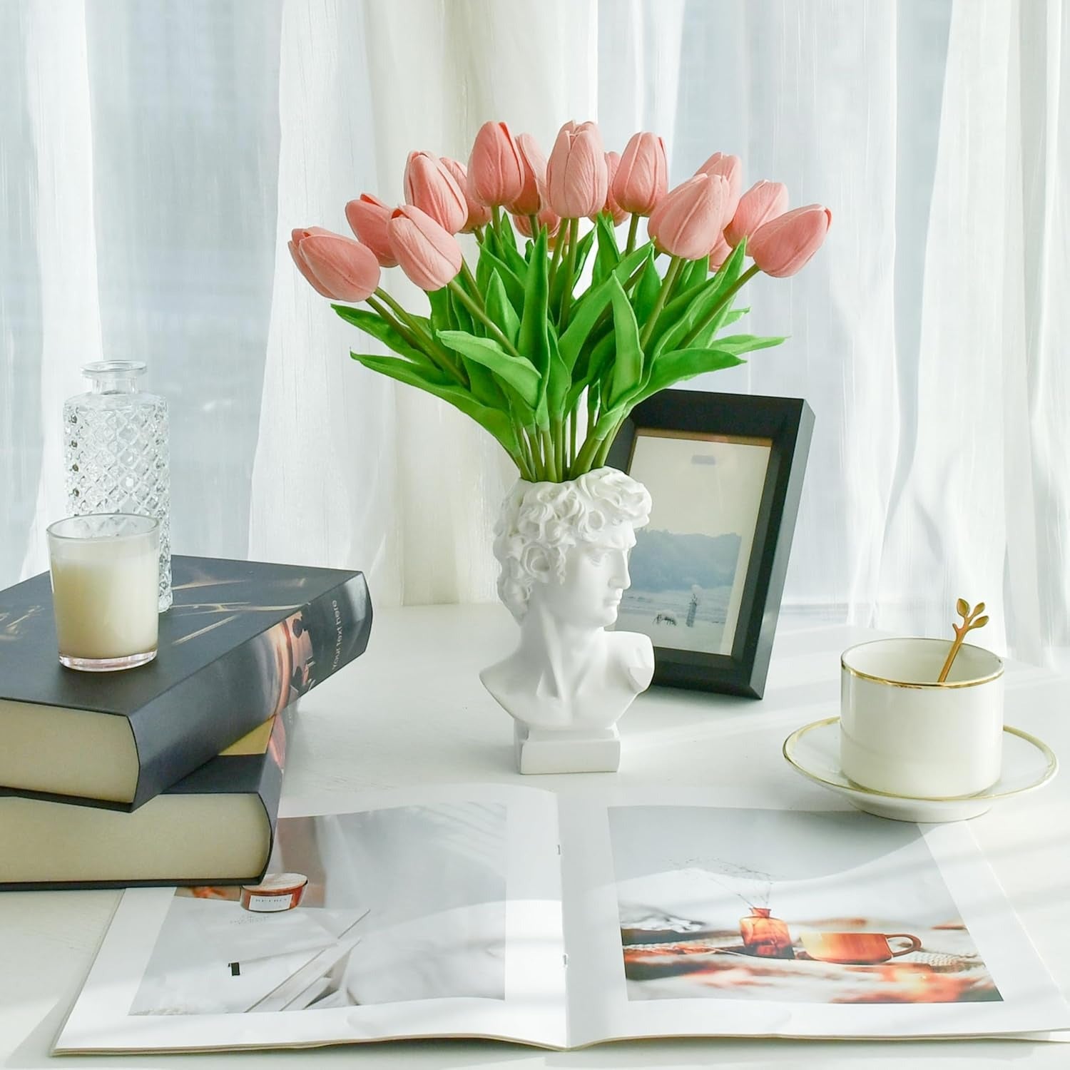 Pink tulips in a white vase on a table with books, a cup, and a candle.