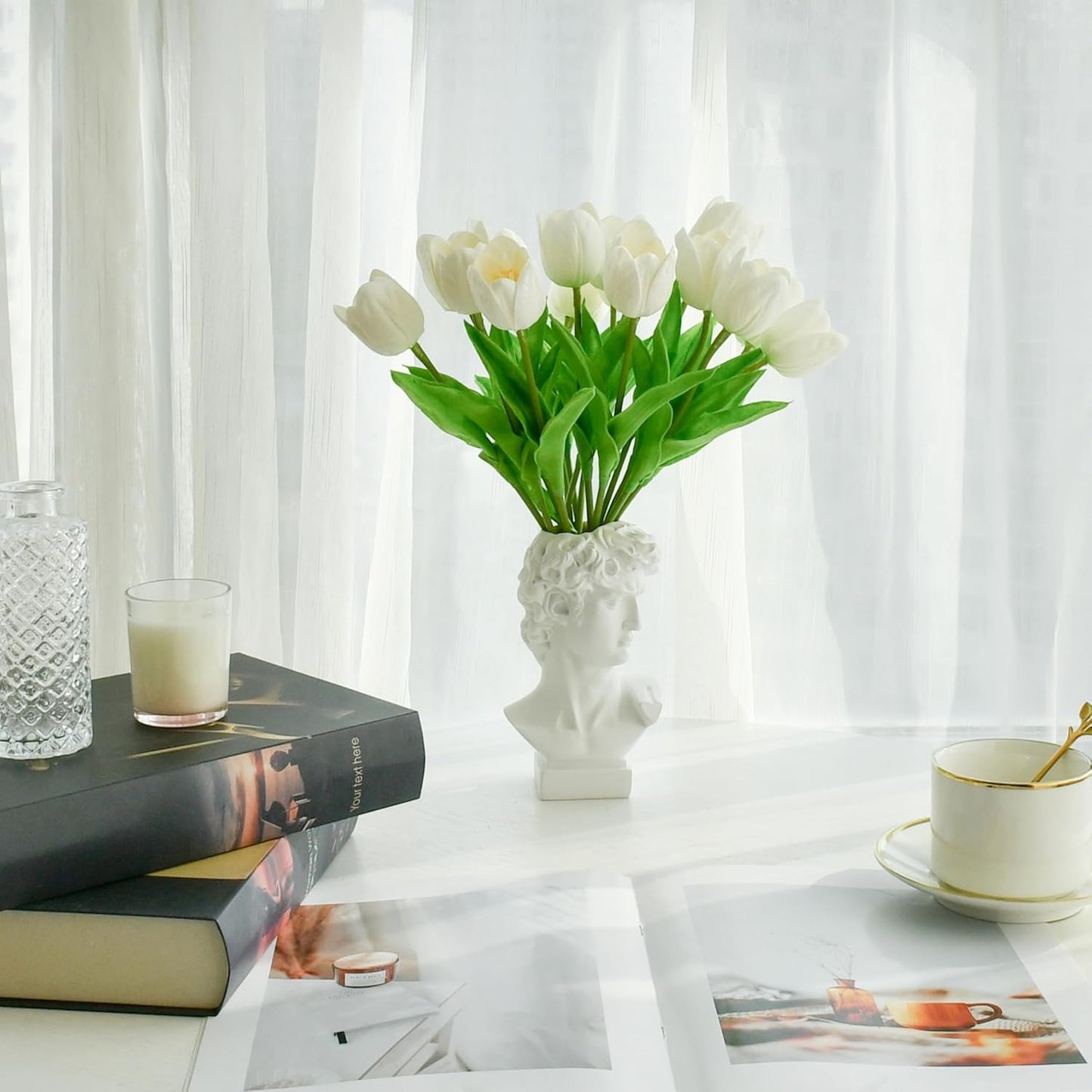 White tulips in a decorative vase on a table with books and a cup.