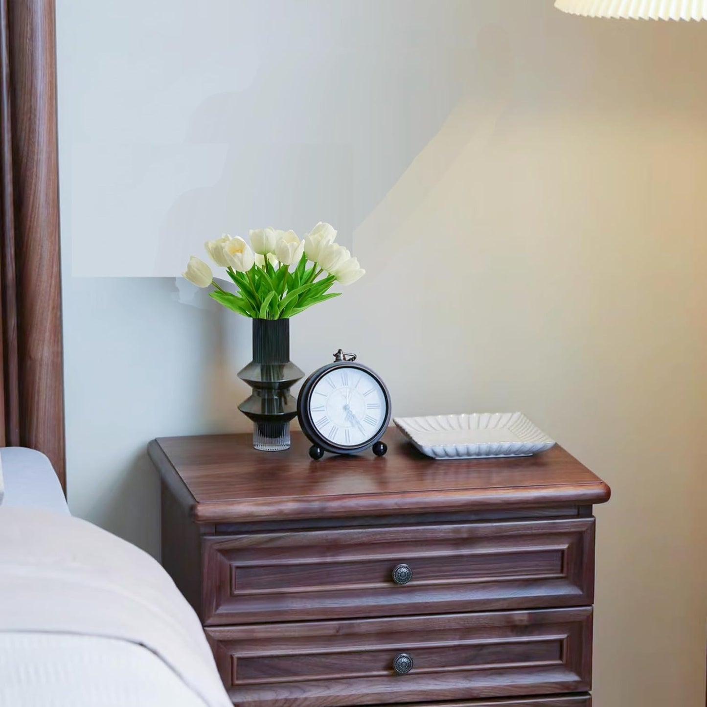 Wooden nightstand with a clock, vase of flowers, and square plate on a blurred bedroom background