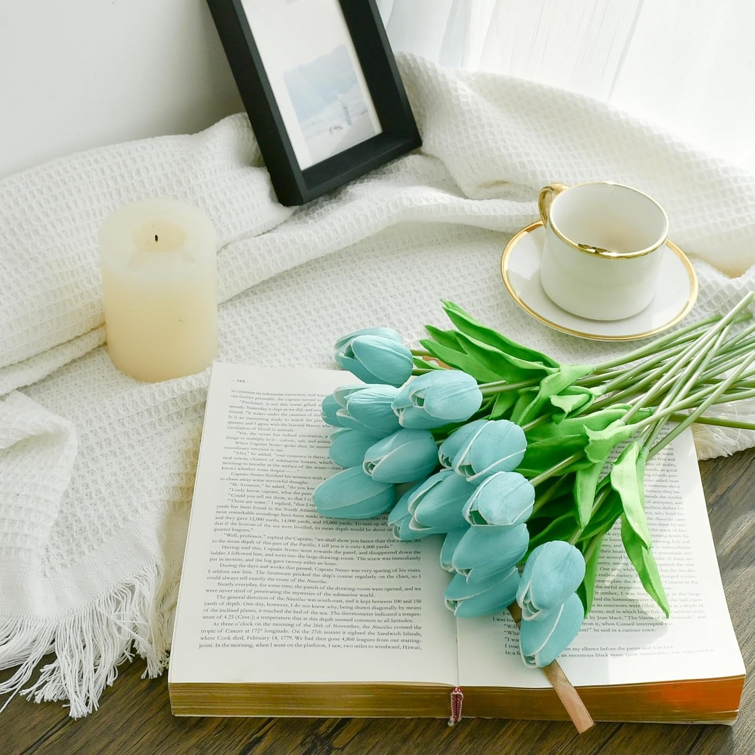 Light blue tulips on an open book with a cup, saucer, and candle in the background.
